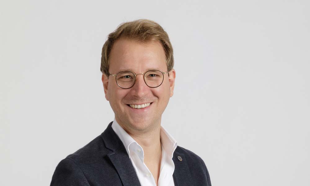 Porträt eines lächelnden Mannes mit Brille und dunklem Blazer vor neutralem Hintergrund.