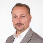 Porträt eines Mannes im hellen Sakko und weißem Hemd, der vor einem neutralen Hintergrund in die Kamera blickt.