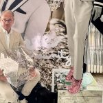 Ein Mann in hellem Anzug sitzt vor einer Modepräsentation und hält einen transparenten Würfel mit dem Wilvorst-Logo in den Händen.