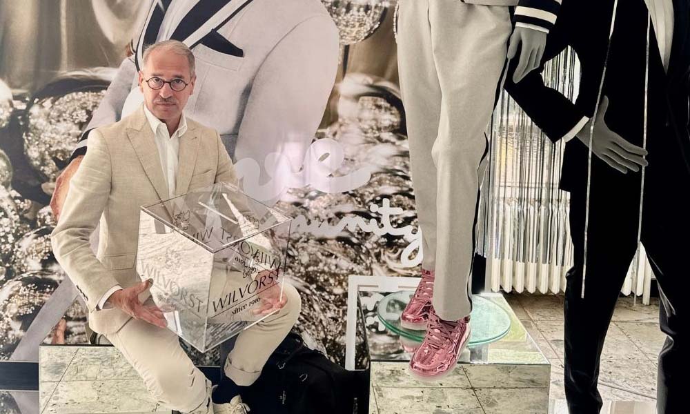 Ein Mann in hellem Anzug sitzt vor einer Modepräsentation und hält einen transparenten Würfel mit dem Wilvorst-Logo in den Händen.