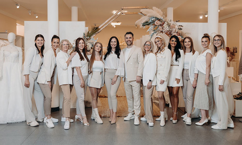 Team eines Brautmodengeschäfts posiert gemeinsam in eleganten, cremefarbenen Outfits zwischen Brautkleidern und moderner Hochzeitsdekoration.