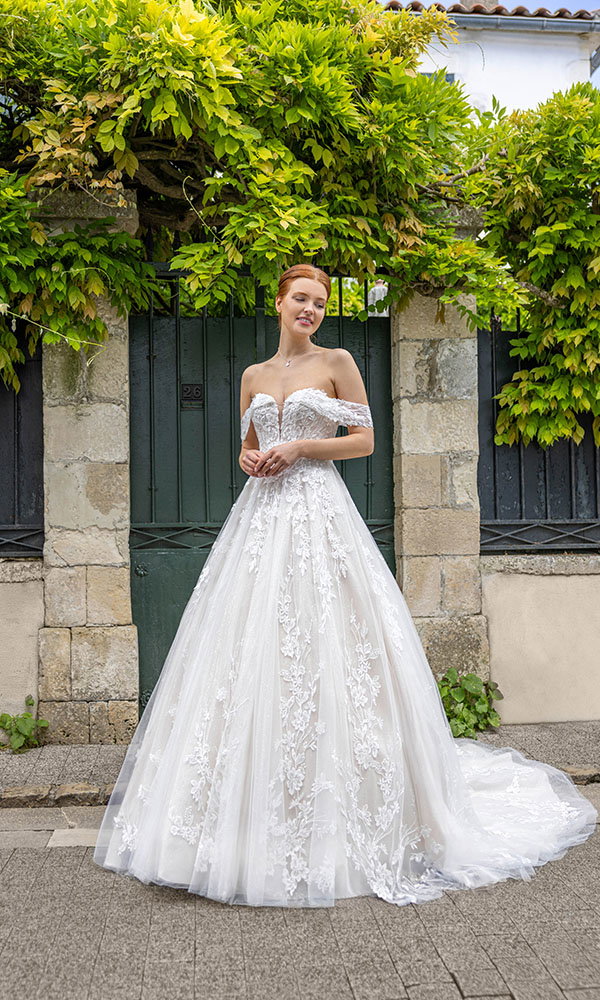 Braut in schulterfreiem Spitzenbrautkleid steht vor einem grünen Tor mit üppiger Pflanzenkulisse.