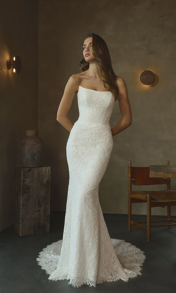 Braut in figurbetontem, trägerlosem Brautkleid steht in stilvollem Interieur mit warmem Licht und minimalistischer Einrichtung.