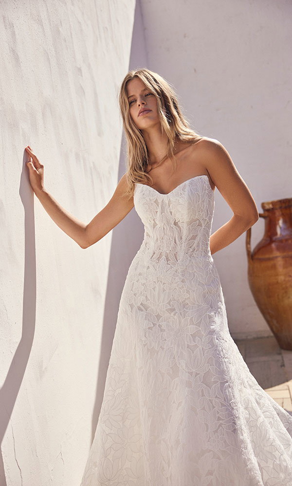 Braut in trägerlosem Spitzenbrautkleid lehnt an einer hellen Wand im Sonnenlicht neben einer Terrakotta-Vase.