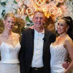 Dick van Zutphen mit Models in Modeca-Brautkleidern vor floraler Fotowand mit leuchtendem Logo auf einem Bridal-Event