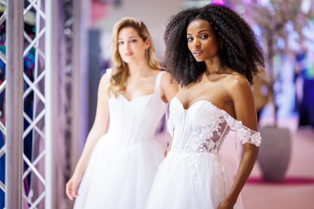 Brautmodel mit natürlicher Lockenpracht trägt ein schulterfreies, detailreich besticktes Brautkleid mit transparenter Corsage, während im Hintergrund ein weiteres Model in einem schlichten weißen Kleid über den Laufsteg schreitet.