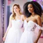 Brautmodel mit natürlicher Lockenpracht trägt ein schulterfreies, detailreich besticktes Brautkleid mit transparenter Corsage, während im Hintergrund ein weiteres Model in einem schlichten weißen Kleid über den Laufsteg schreitet.