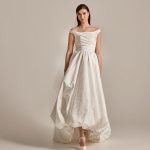 Braut in minimalistischem Off-Shoulder-Brautkleid mit voluminösem Rock steht vor neutralem Hintergrund.