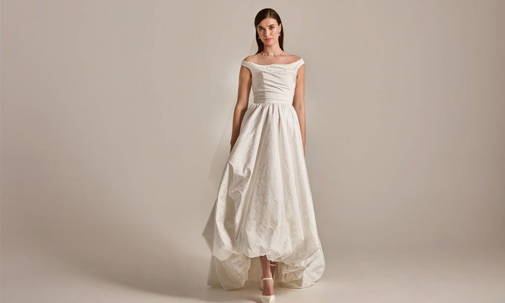 Braut in minimalistischem Off-Shoulder-Brautkleid mit voluminösem Rock steht vor neutralem Hintergrund.