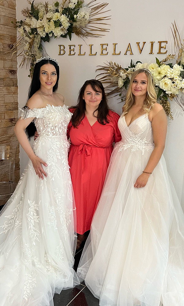 Zwei Bräute in eleganten Spitzen-Brautkleidern stehen neben einer Frau im roten Kleid vor einer dekorativen Wand mit der Aufschrift Bellelavie.