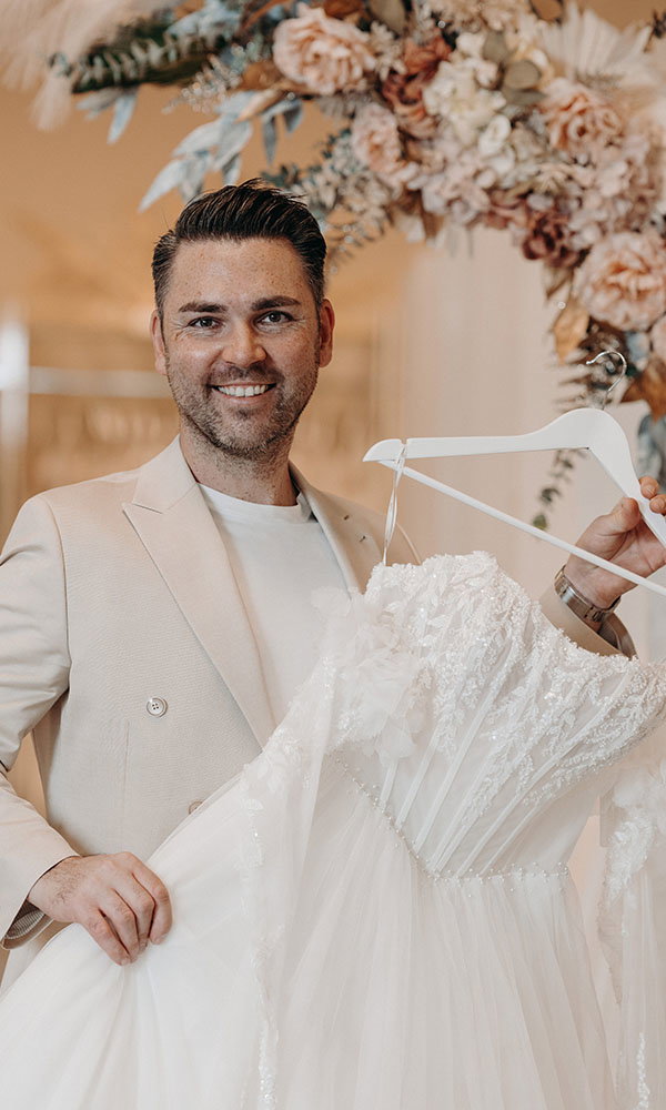 Mann in hellem Anzug hält lächelnd ein Brautkleid auf einem Bügel in einem stilvollen Brautmodengeschäft.