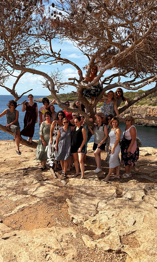 Gruppe von Frauen posiert unter einem knorrigen Baum an einer felsigen Küste mit Blick auf das Meer.