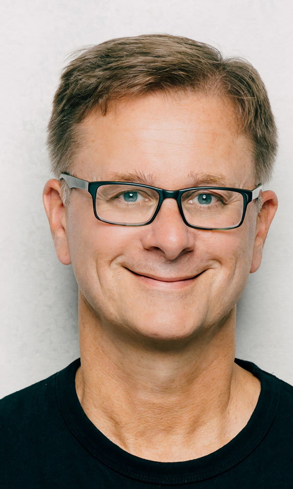 Porträt eines lächelnden Mannes mit kurzer Frisur und Brille vor neutralem Hintergrund.
