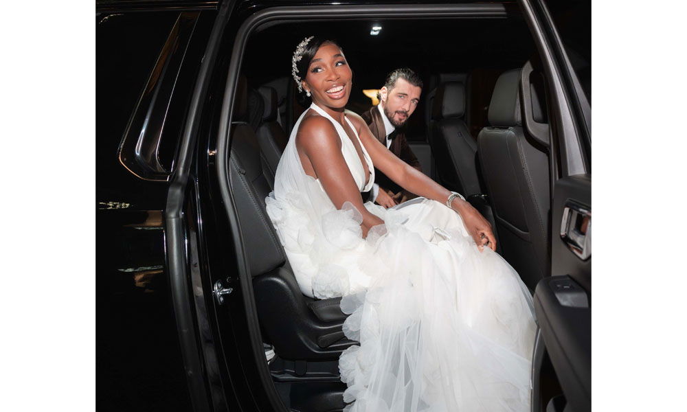 Braut im weißen, voluminösen Tüllkleid mit glitzerndem Haaraccessoire steigt lächelnd aus einem schwarzen Auto, während ihr Partner im Hintergrund im Fahrzeug sitzt.