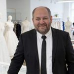 Mann im schwarzen Anzug steht lächelnd in einem Brautmodengeschäft, umgeben von Schaufensterpuppen mit verschiedenen Brautkleidern und Stoffen auf einem Arbeitstisch.