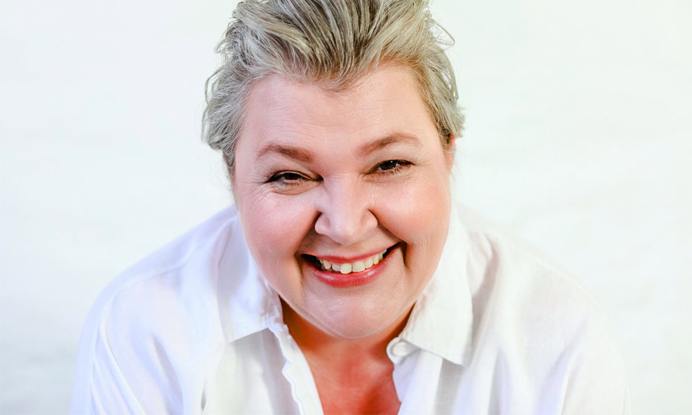 Porträt einer lächelnden Frau mit kurzen grauen Haaren und weißer Bluse, die freundlich und offen in die Kamera blickt, vor hellem, neutralem Hintergrund.