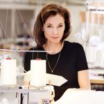 Frau arbeitet an einer Nähmaschine in einem Atelier und blickt direkt in die Kamera, umgeben von Stoffen und Garnrollen.