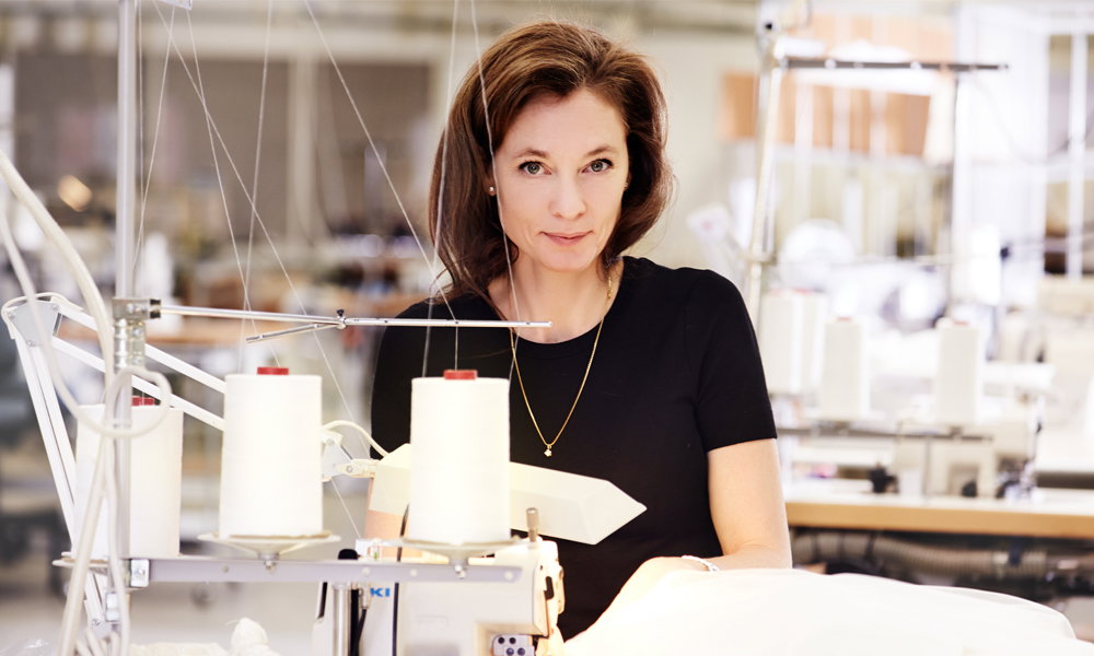 Frau arbeitet an einer Nähmaschine in einem Atelier und blickt direkt in die Kamera, umgeben von Stoffen und Garnrollen.