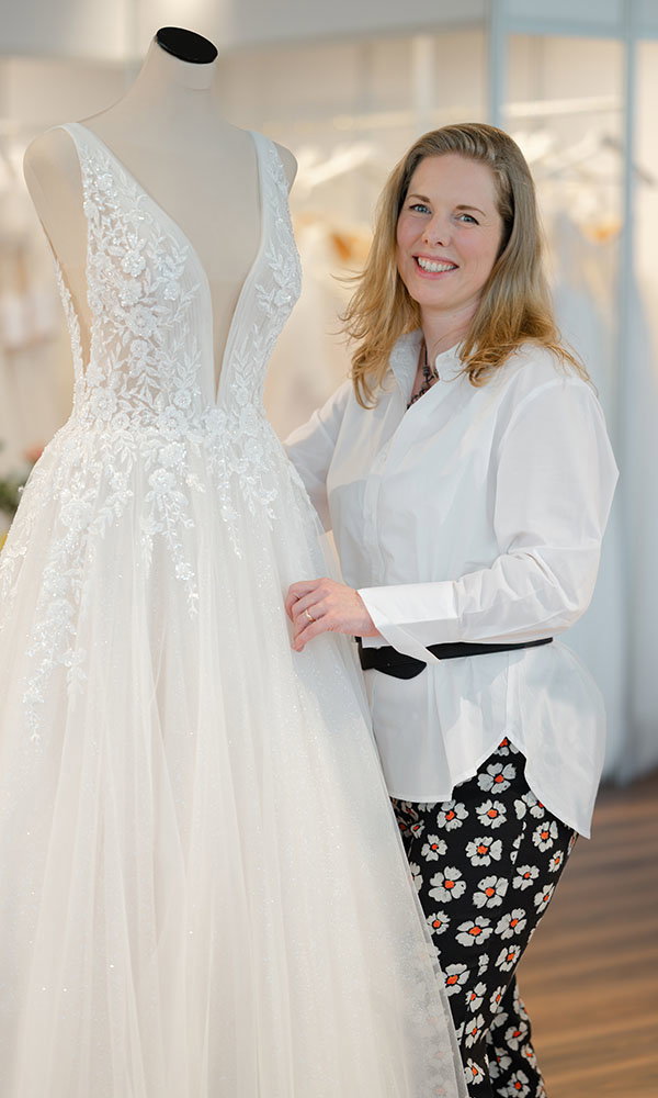 Frau in weißer Bluse steht neben einem Brautkleid mit Spitze und hält den Stoff, während sie in einem hellen Atelier lächelt.