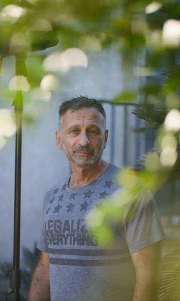 Mann mit grauem T-Shirt steht im Freien, teilweise von unscharfen Blättern umrahmt, und blickt in die Kamera.