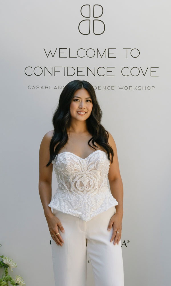 Frau in weißem Brauttop und Hose steht lächelnd vor einer Wand mit der Aufschrift „Welcome to Confidence Cove“.