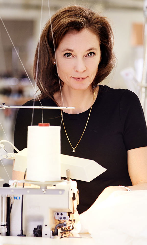 Frau sitzt an einer Nähmaschine im Atelier und blickt in die Kamera, während sie an Stoff arbeitet.