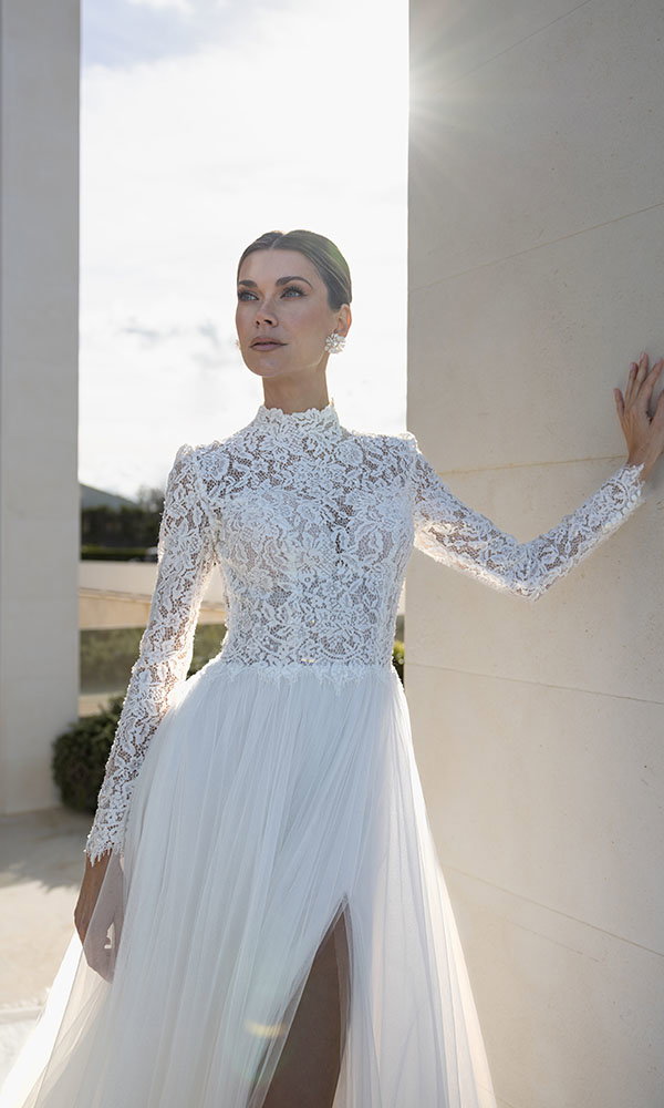 Braut in hochgeschlossenem Spitzenbrautkleid mit langen Ärmeln steht im Sonnenlicht an moderner Architektur.