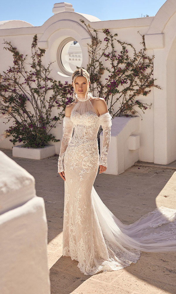 Braut in figurbetontem Spitzenbrautkleid mit langen Ärmeln steht vor weißer Architektur mit blühenden Ranken im Sonnenlicht.