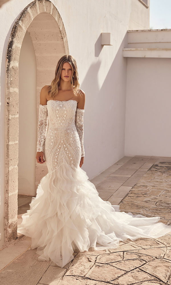 Braut in schulterfreiem Spitzenbrautkleid mit voluminösem Tüllrock steht unter einem Torbogen in mediterranem Ambiente.