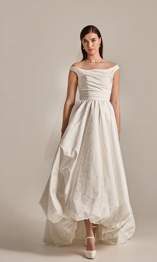 Braut in minimalistischem Off-Shoulder-Brautkleid mit voluminösem Rock steht vor neutralem Hintergrund.