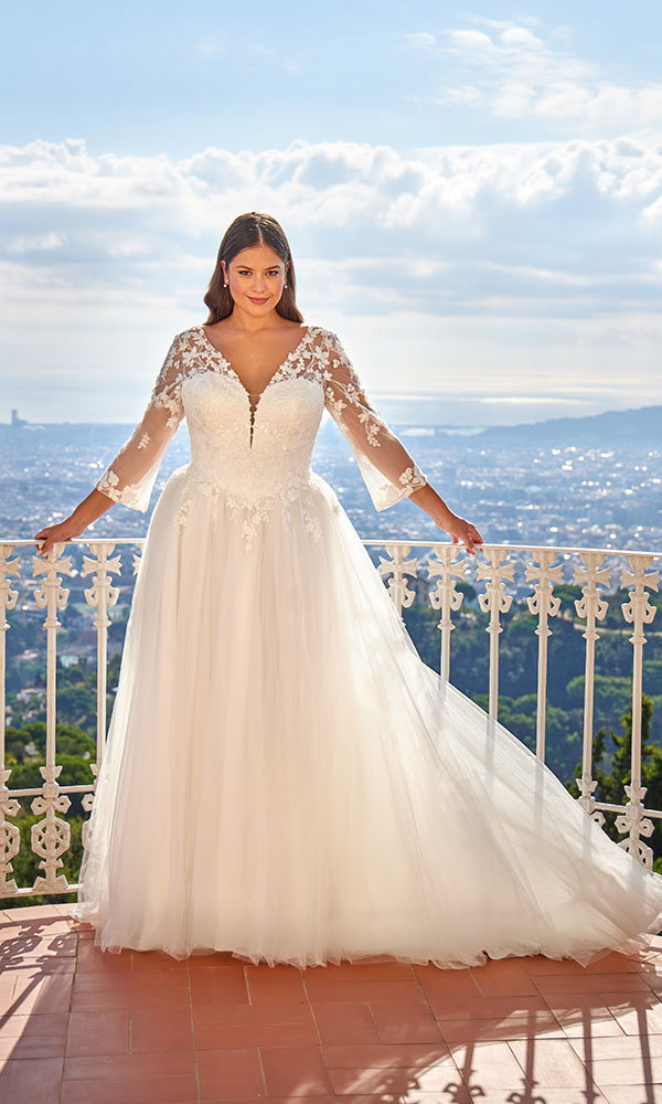 Braut in A-Linien-Brautkleid mit Spitzenärmeln steht auf einem Balkon mit Blick über eine Stadtlandschaft.
