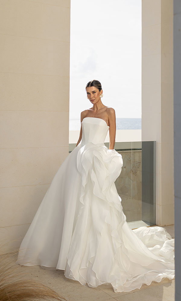 Braut in trägerlosem Brautkleid mit voluminösen Rüschen steht in heller Architektur mit Blick aufs Meer.