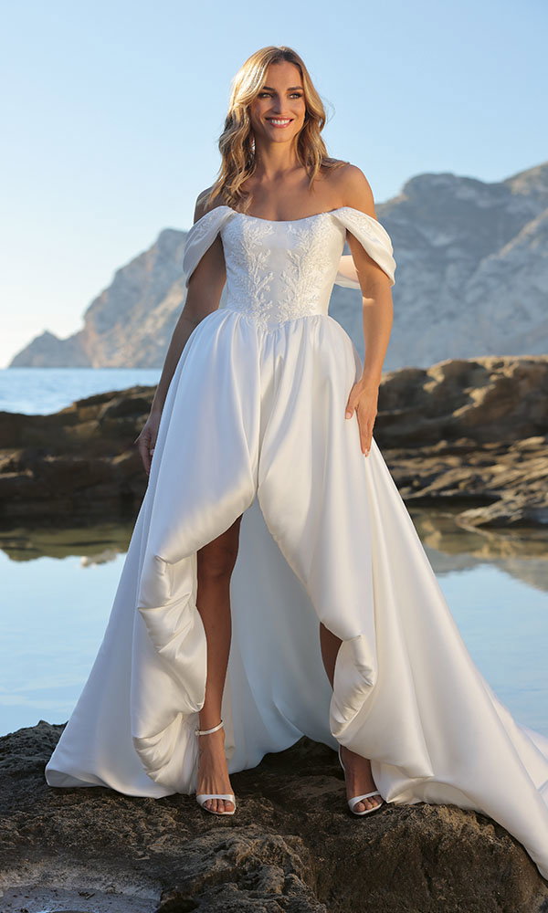 Braut in modernem Brautkleid mit Schlitz und Off-Shoulder-Trägern steht auf Felsen am Meer vor Bergkulisse.