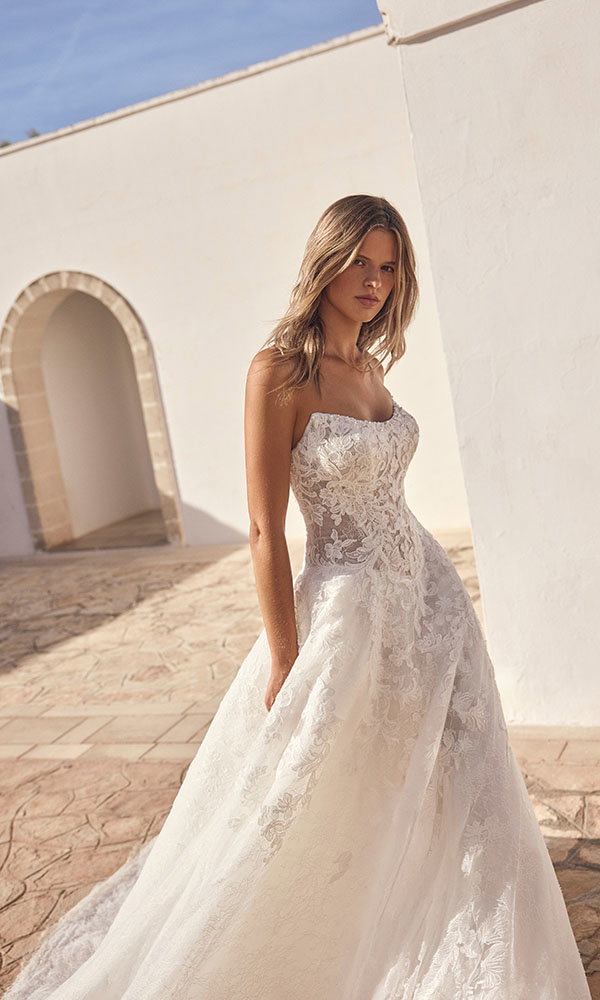 Braut in trägerlosem Spitzenbrautkleid steht in mediterraner Architektur im warmen Sonnenlicht.