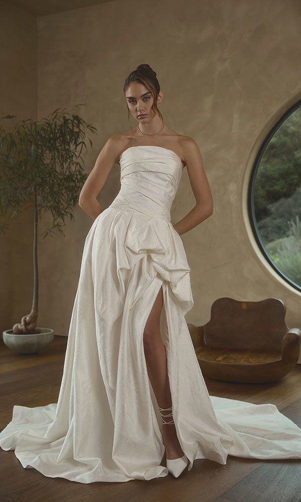 Braut in trägerlosem Brautkleid mit Drapierungen und hohem Schlitz steht in stilvollem Interieur vor rundem Fenster.