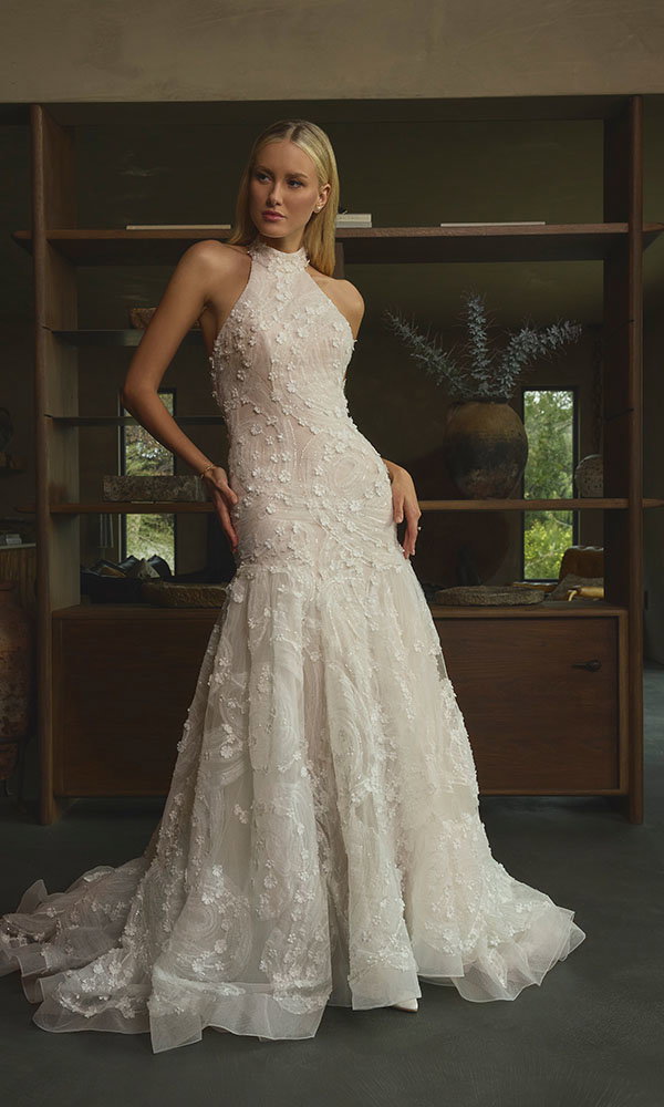 Braut in detailreichem Neckholder-Spitzenbrautkleid steht in stilvollem Interieur vor einem Holzregal.