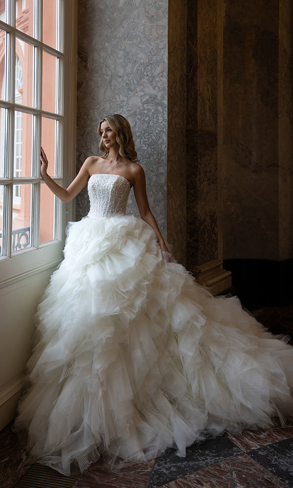 Braut in trägerlosem Brautkleid mit opulentem Tüllrock steht am Fenster in elegantem Interieur.