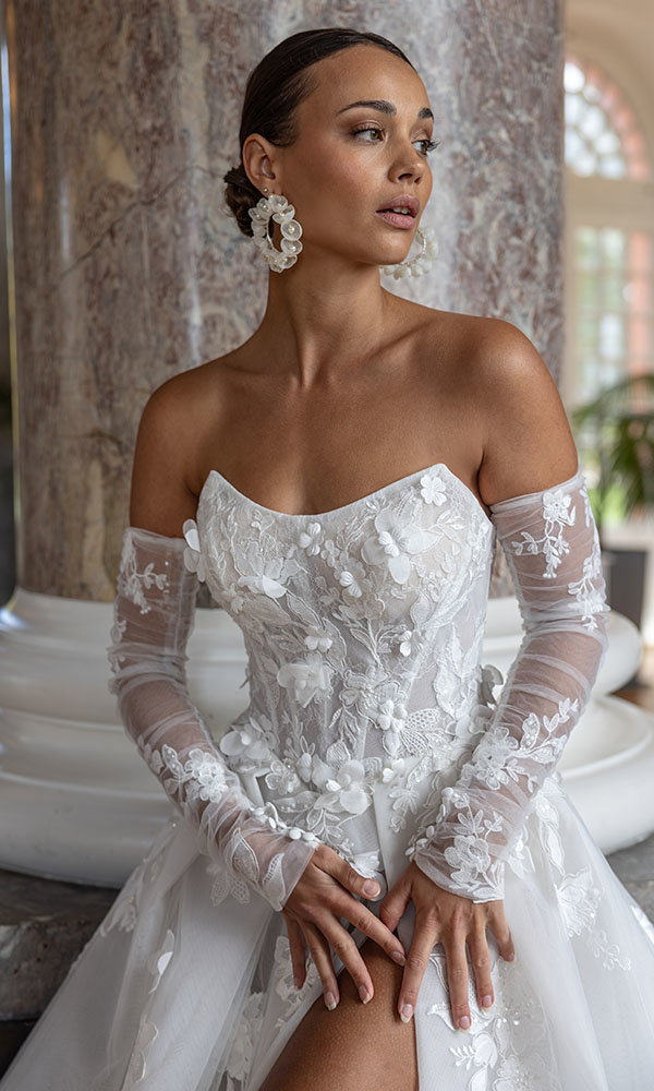 Braut in schulterfreiem Spitzenbrautkleid mit transparenten Ärmeln sitzt vor einer Marmorsäule und blickt zur Seite.