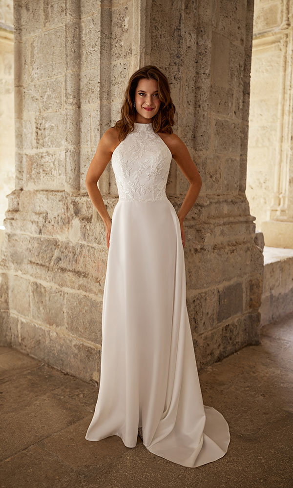 Braut in schlichtem Neckholder-Brautkleid steht vor steinerner Säule in historischem Ambiente.