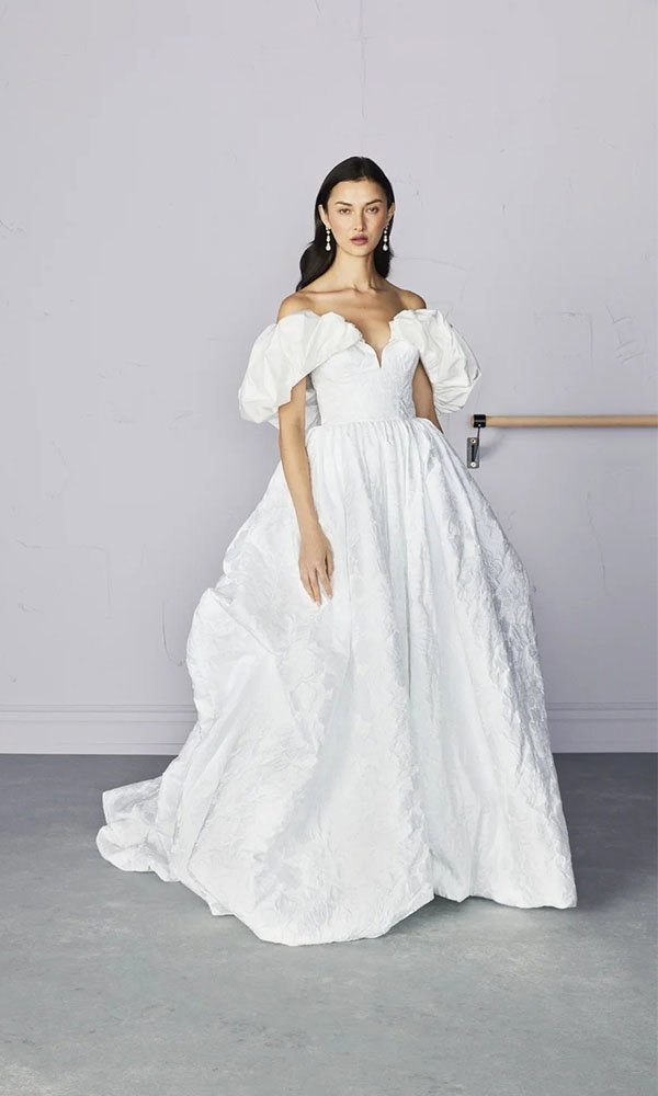 Braut in schulterfreiem Brautkleid mit voluminösen Puffärmeln steht vor schlichtem Hintergrund im Studio.